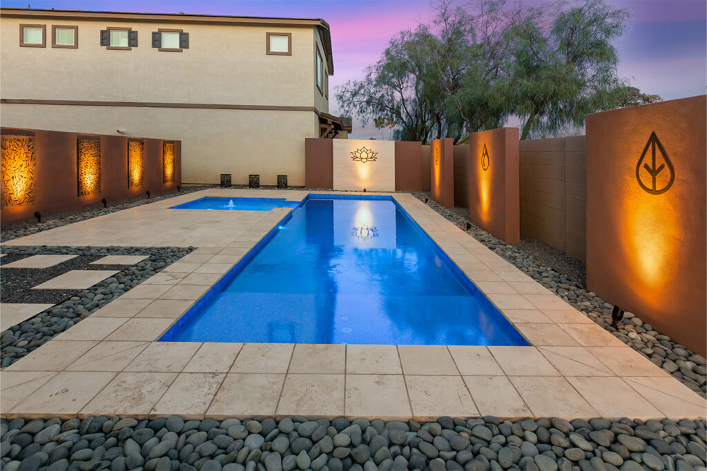Modern backyard pool in Phoenix with travertine tile, illuminated wall art, and decorative stone landscaping.