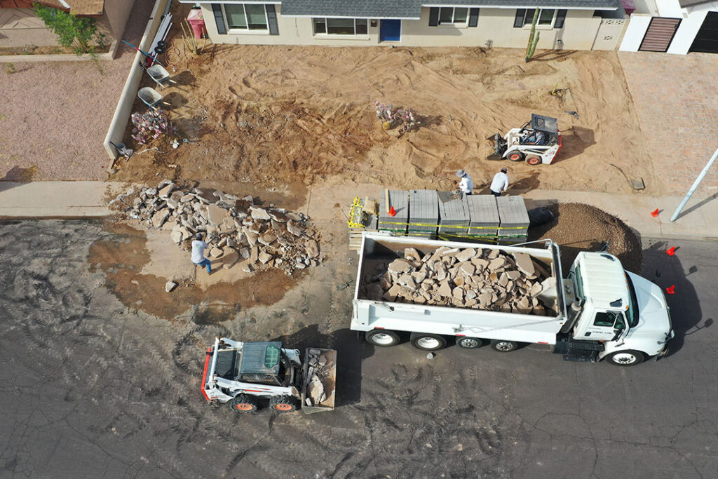 Crew preparing a Phoenix front yard for paver installation with heavy equipment and demolition debris.
